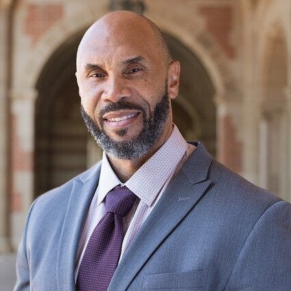 Darnell Hunt, a Black man with a shaved head and beard, smiles for a portrait wearing a suit.