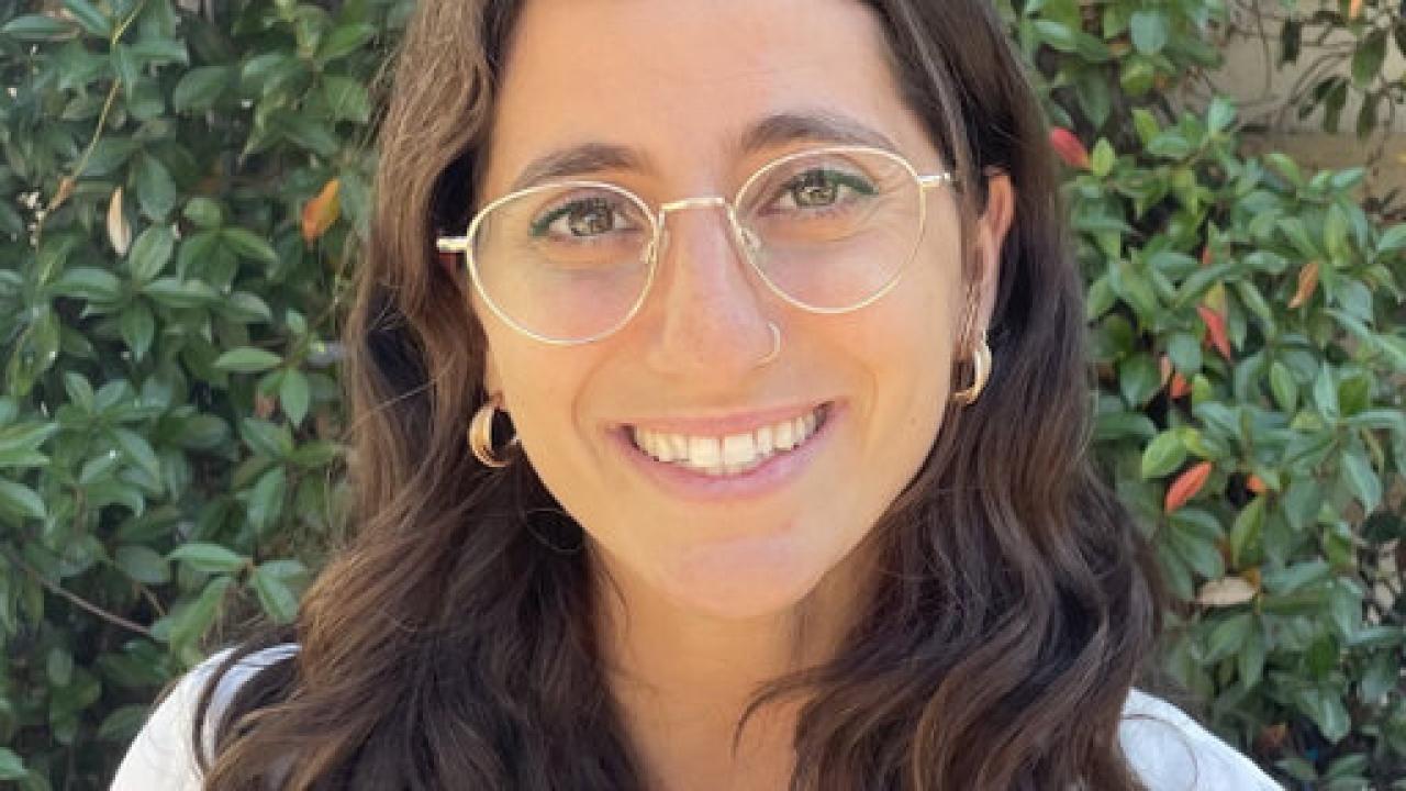 A woman with an olive skin tone and wavy, shoulder-length brown hair smiles.