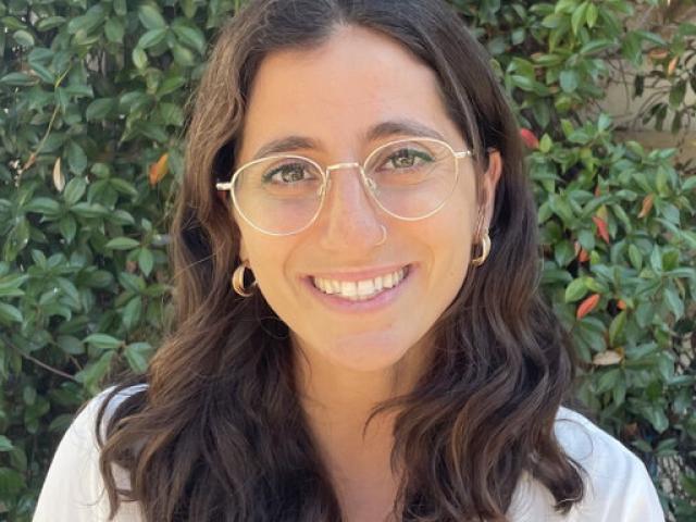 A woman with an olive skin tone and wavy, shoulder-length brown hair smiles.