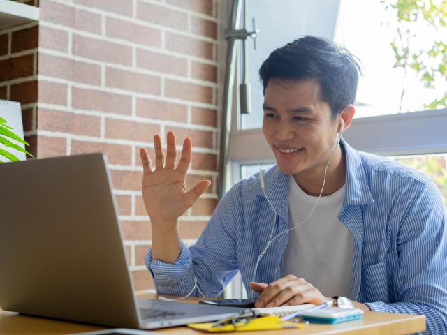 A young man waves at someone on a laptop.