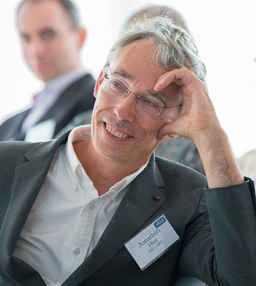 Jonathan Flint leans his head on his hand. He has light skin, gray hair, glasses, and is wearing a gray suit.