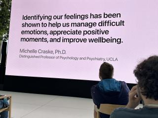 A large LED screen at a talk displays a quote "Identifying our feelings has been shown to help us manage difficult emotions, appreciate positive moments, and improve wellbeing." attributed to Michelle Craske, PhD, Distinguished Professor of Psychology and Psychiatry, UCLA